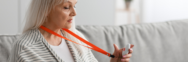 photograph of a woman with white hair holding a health alarm pendant. She has an orange lanyard around her neck.